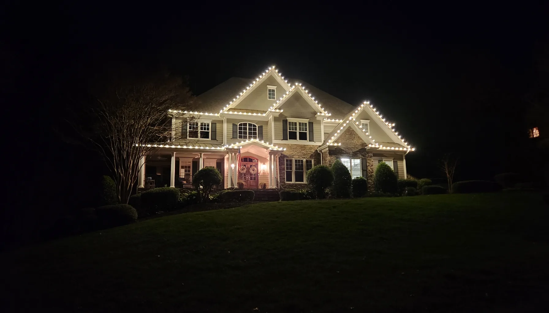 Two-story home near Sunset Lake in Holly Springs, NC with warm white C9 Christmas lights outlining every roofline and gable, and lit wreaths on the front door